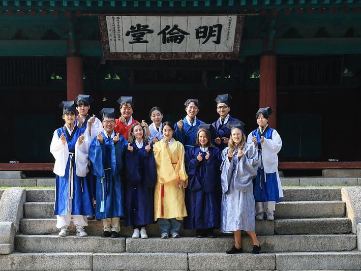 Twelve individuals dressed in traditional Korean scholar Hanbok smile for a photo.