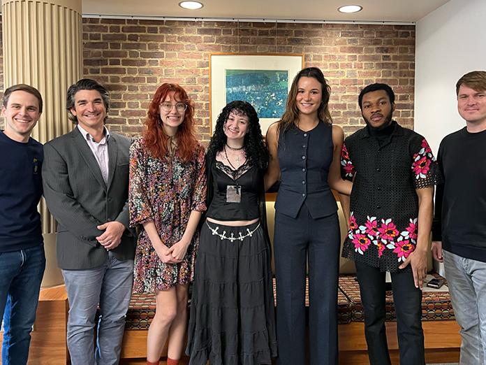 Group photo of seven people form left a white man with brown hair, a white man with grey and black hair, a white young woman with red hair, a white young woman with black hair, a white young woman with brown hair, a black man with dark hair and a white man with brown hair