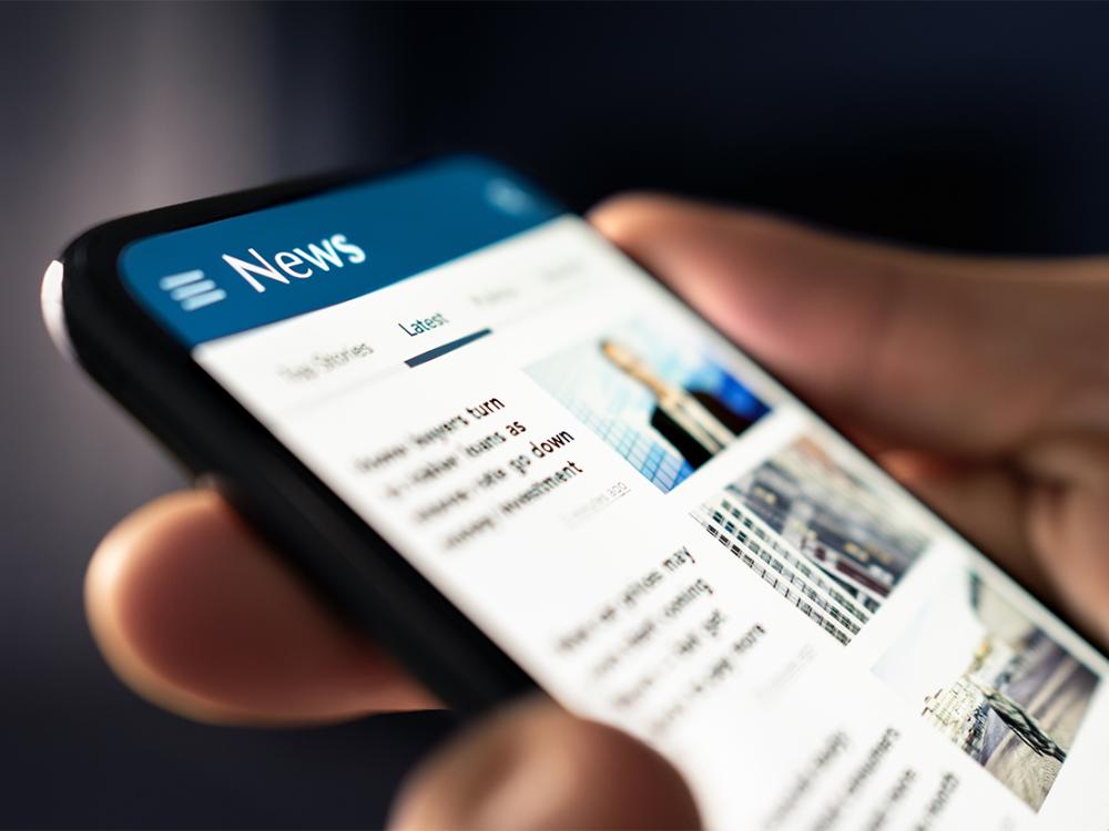 A hand holding a mobile device with a newsfeed in view.