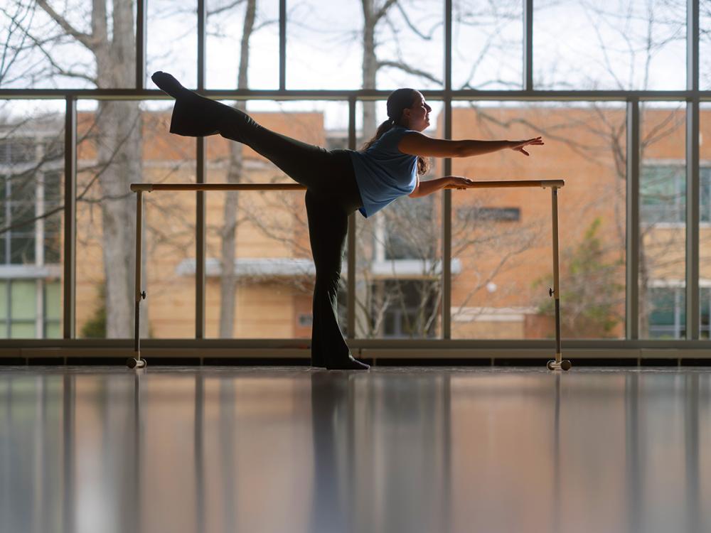 Dancer practices at a ballet barre with one leg extended behind in a studio with large windows and reflected floor light