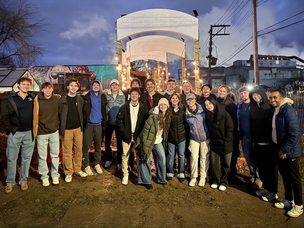 Group of people standing together at dusk beneath illuminated wooden archways along an outdoor garden path, with city buildings and power lines in the background.