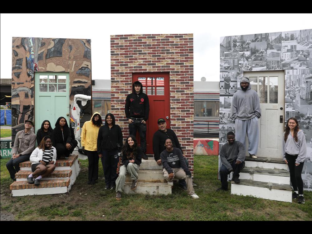 Group of people pose on steps in front of three small facade panels—one with a green door and mural, one with a red door and brick pattern, and one with a white door and black‑and‑white collage—set outdoors on grass.