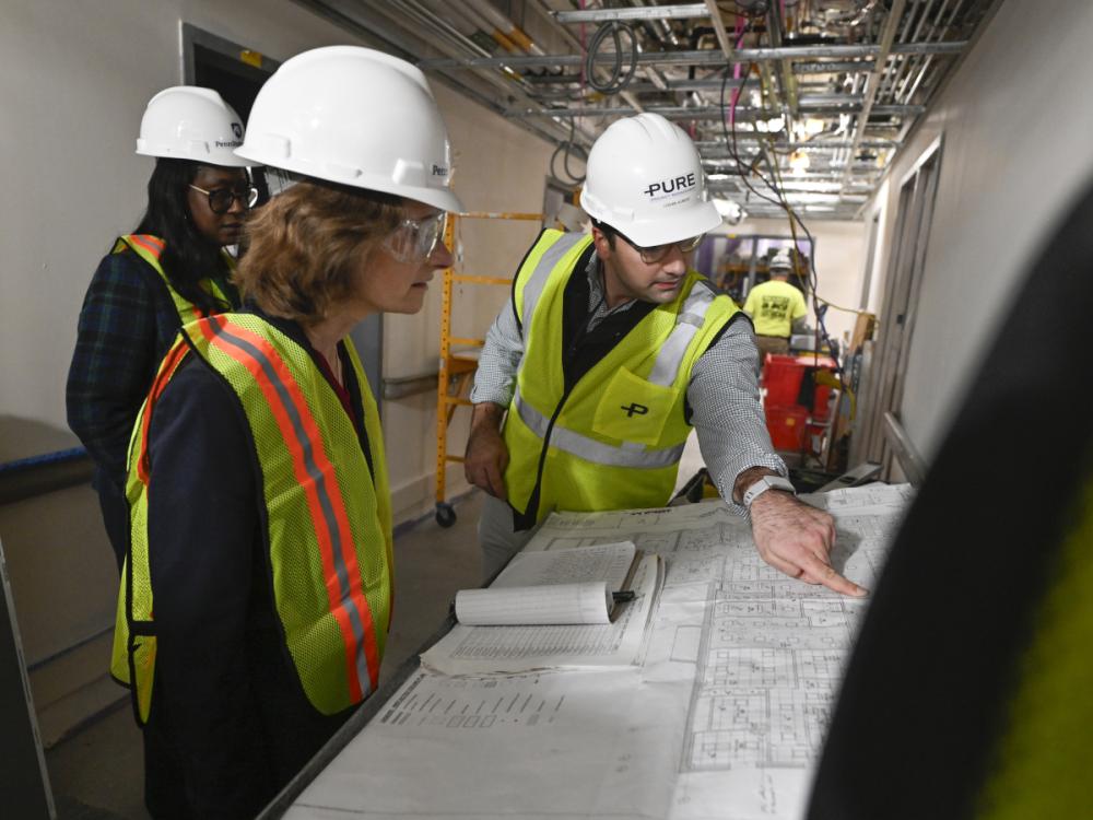 A man in a hard hat points to blueprints laid out on a table while two women in hard hats look on. They’re standing in a hallway that’s under construction.