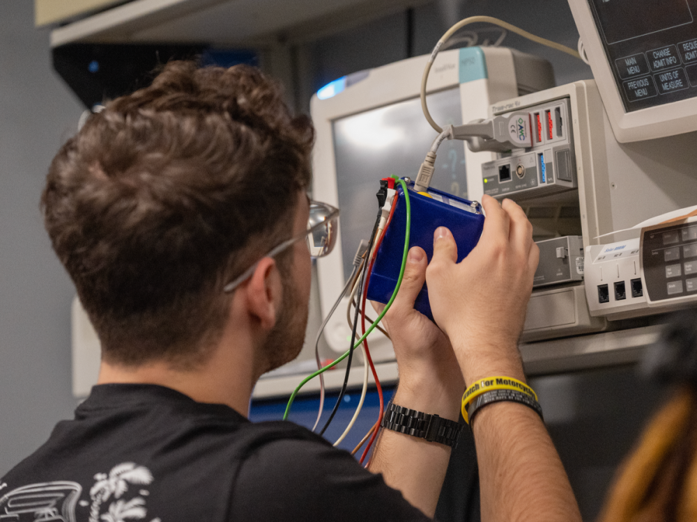 A student works on biomedical equipment in a laboratory setting, connecting diagnostic cables and testing devices attached to a hospital-style patient monitor and medical equipment system.
