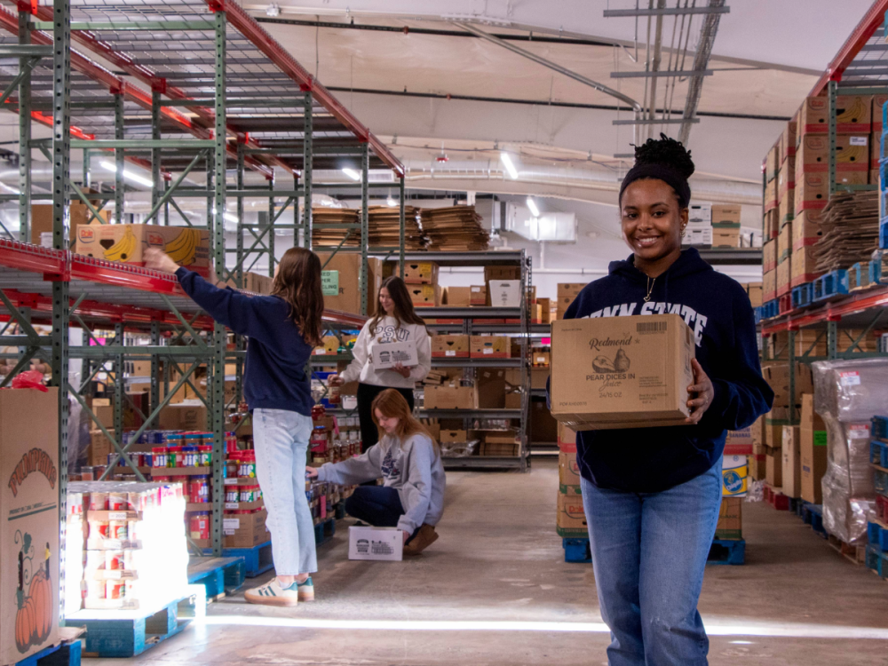 A young woman holds a cardboard box while standing in a warehouse space full of shelves stacked with food products.  Three other people stack food on the shelves behind her.