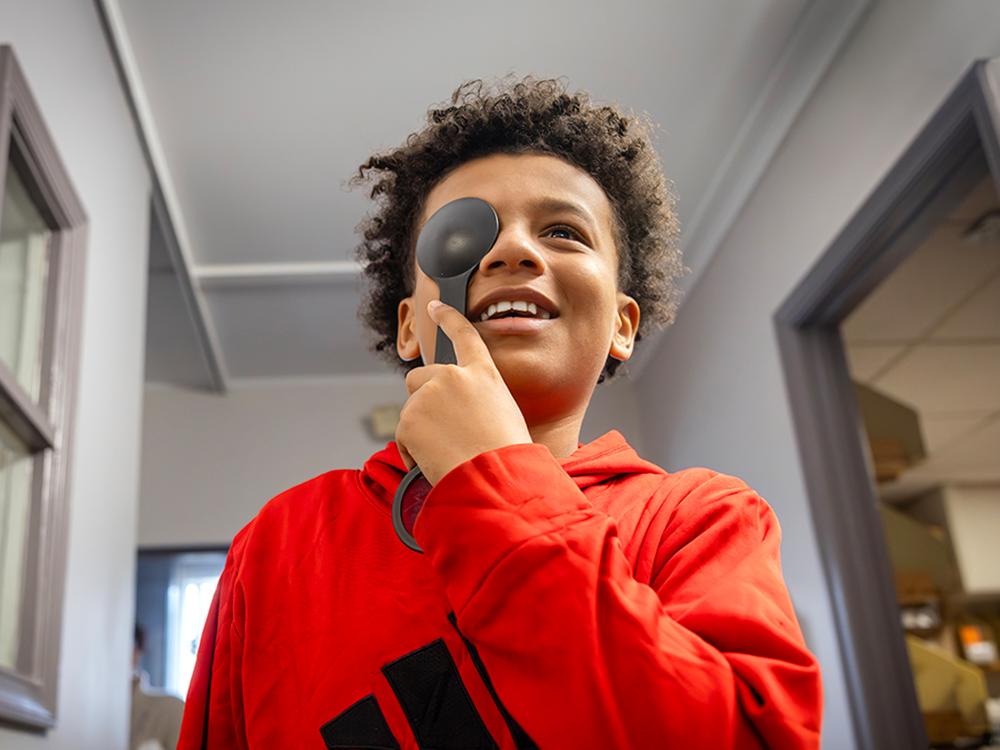 A boy holds an occluder over his right eye and smiles as he looks at a vision chart. He is wearing a sweatshirt with an Adidas logo on it.