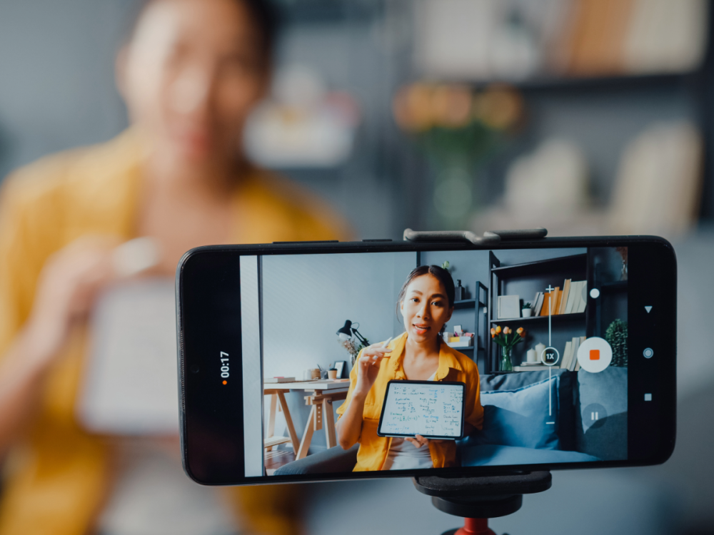 photo of iphone on tripod recording woman making educational video holding e-tablet and pointing to content on screen