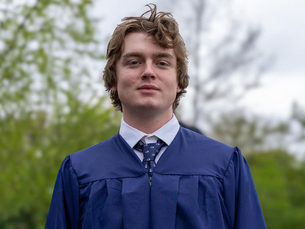 Sailor Walter in a blue graduation gown and tie stands outdoors. The background features blurred green foliage and a cloudy sky.