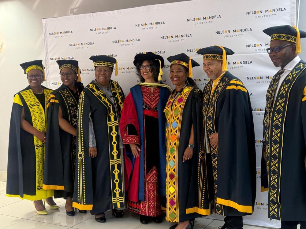Gabeba Baderoon wears colorful regalia while standing in the middle of a group of other people wearing academic regalia.
