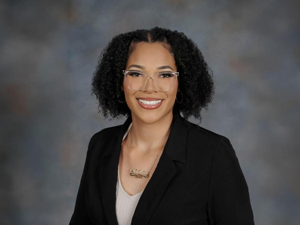 Christina Luisa Hughes, with curly hair, glasses, and a black blazer smiles in a professional studio portrait with a neutral gradient background.