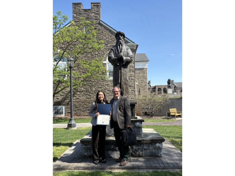A student and college professor stand together holding and award in front of a statue on a campus
