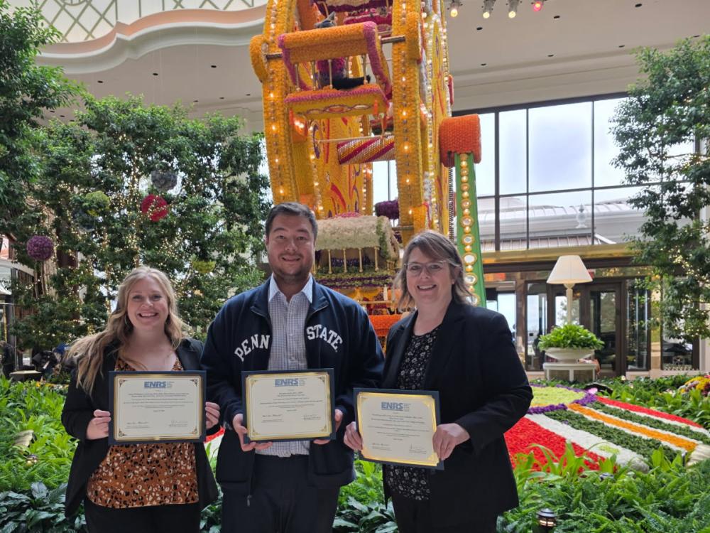 Three people standing indoors holding certificates with a large colorful floral Ferris wheel sculpture and lush greenery in the background.