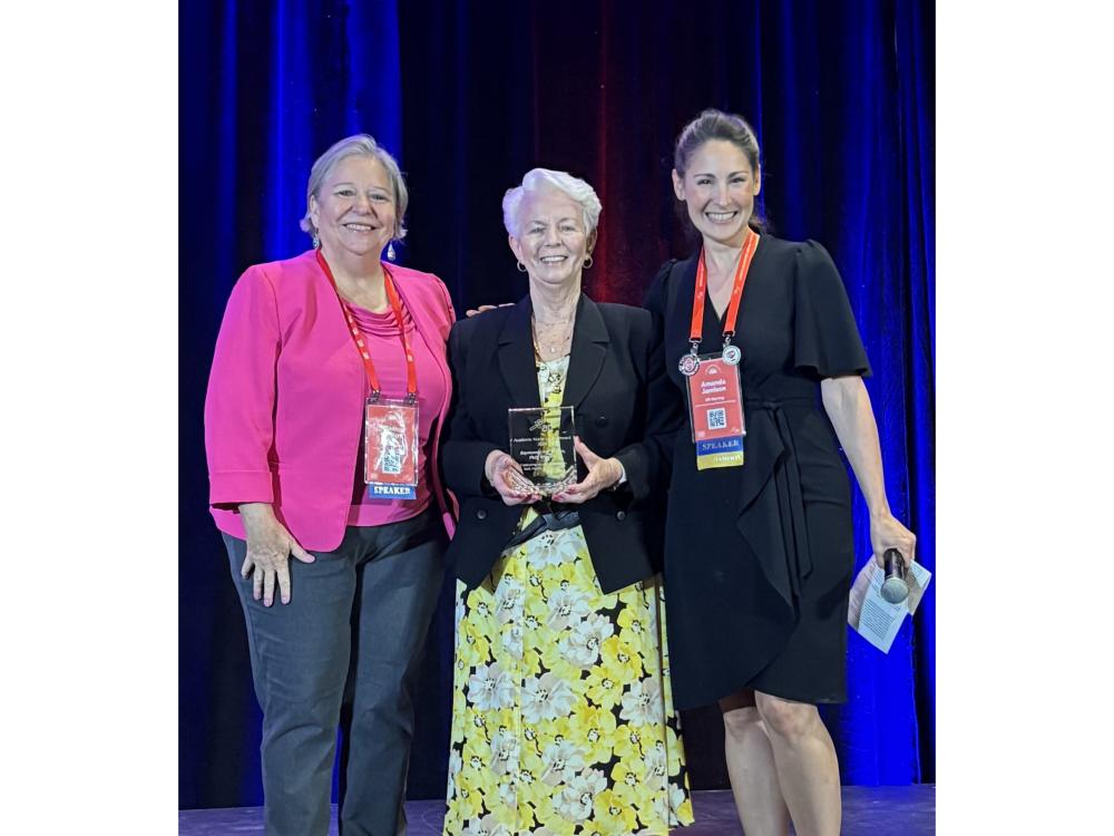 Three women stand on stage at an award ceremony. The woman in the center, Rae Brown, holds a glass award.