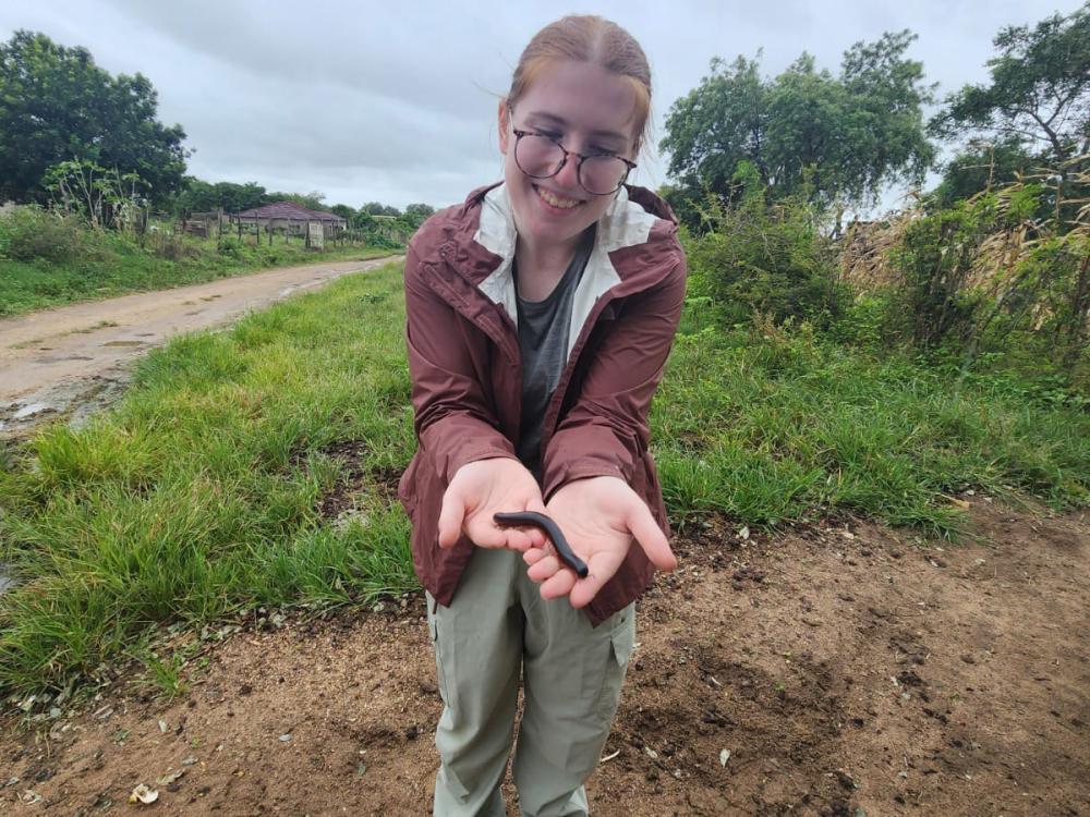 Isabel Rivera wearing glasses and a brown jacket stands on a dirt path, holding a small black creature in their hands and smiling. Greenery and cloudy sky are in the background.