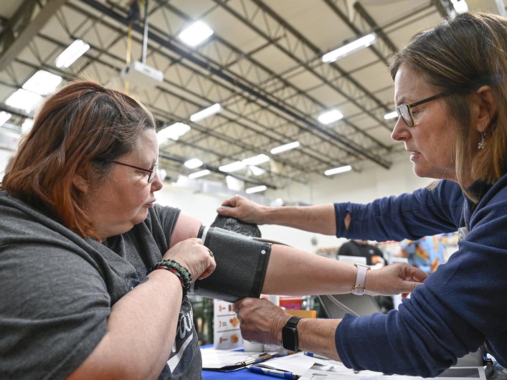 A woman holds a blood pressure cuff on her arm as a nurse on the right adjusts it. They are sitting at a table in a large warehouse room with a high ceiling and bright fluorescent lights.