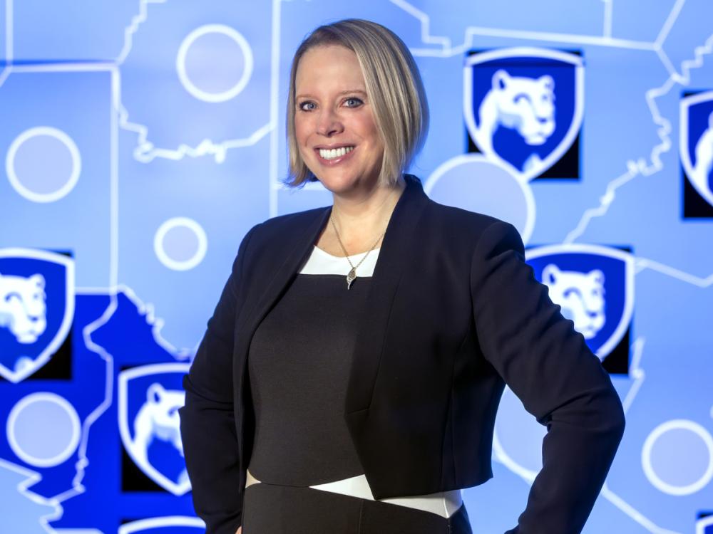 Jennifer Kraschnewski poses for a photograph, wearing professional attire. The background is a map with several Penn State Nittany Lion shield logos.