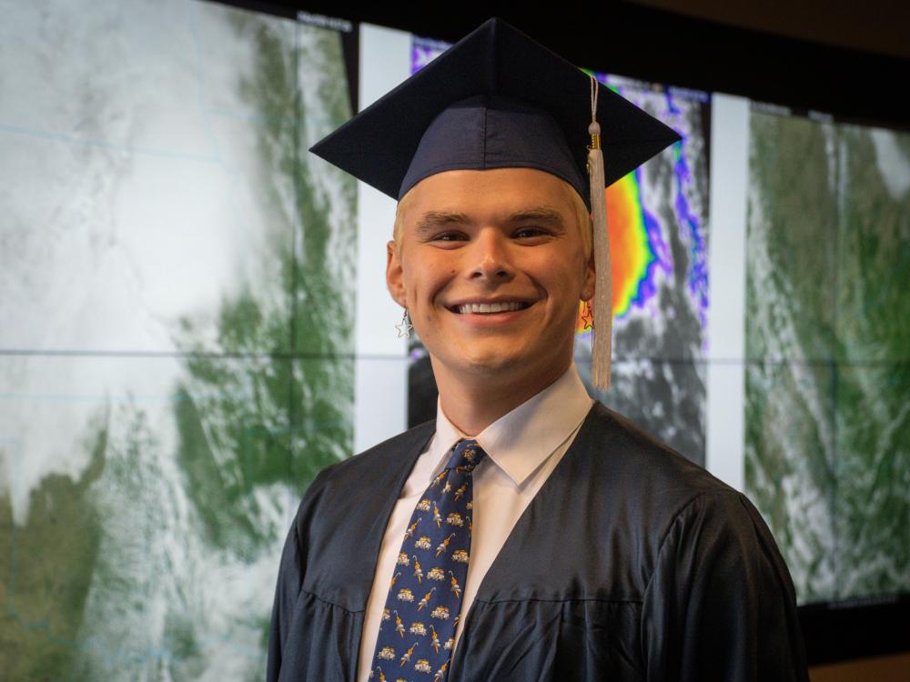 Person smiling, wearing a graduation cap and sitting in front of a weather wall