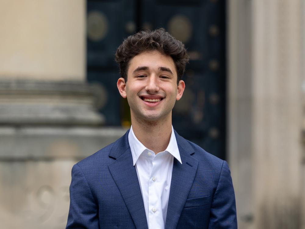 Max wears a blue suit and white shirt while standing in front of Sparks Building.