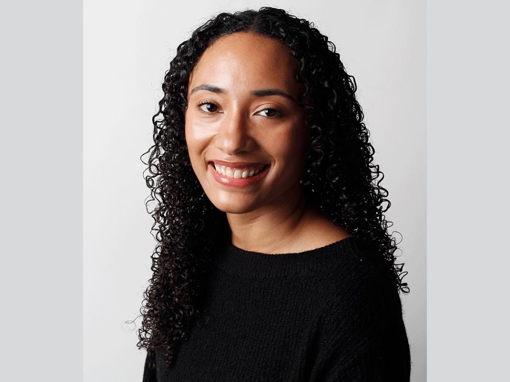 Studio portrait of a person with long, tightly curled hair, facing slightly to the right and looking toward the camera. The person is wearing a black sweater and has a neutral facial expression with teeth visible. The background is a plain light gray.