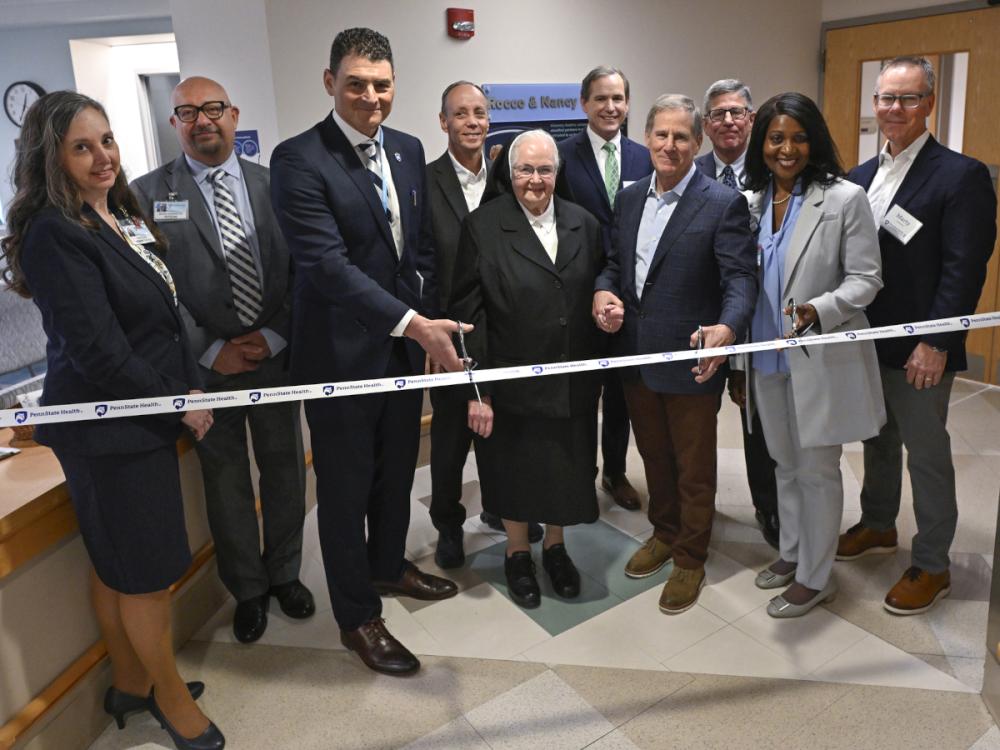 Ten people wearing professional attire pose for a photo in a hospital lobby area as they prepare to cut a ribbon with the “Penn State Health” logo.