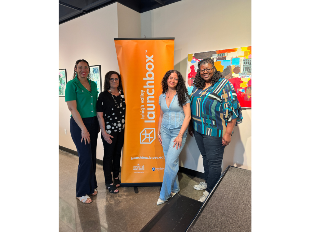 four women smile in front of an orange launchbox banner inside an art gallery