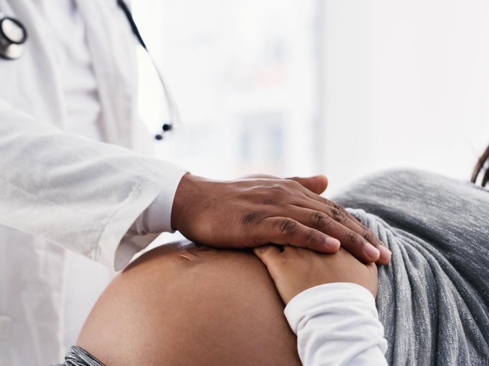 During a visit to a doctor’s office, a doctor and a pregnant person rest their hands on the baby bump. Their faces are not visible.