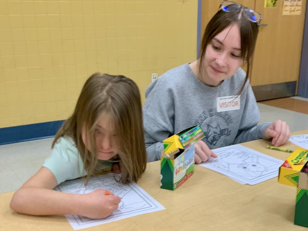 A young student sits at a table and colors while a Penn College student watches.