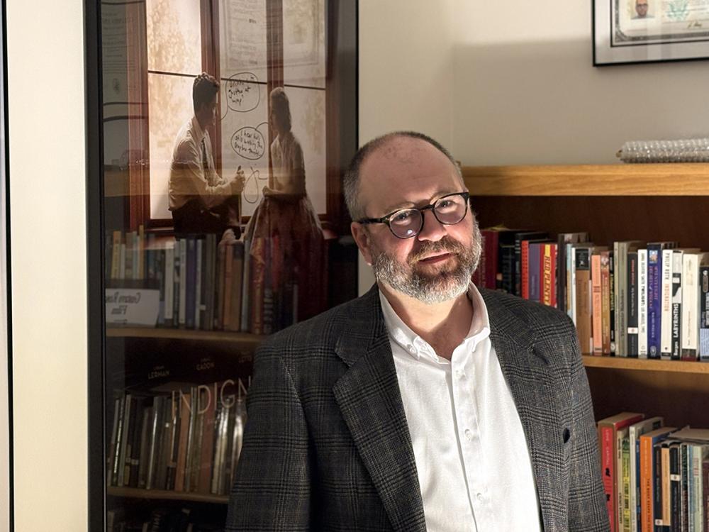 A man in a sports jacket stands in a dimly lit office with movie posters on the wall and a bookshelf in the background.