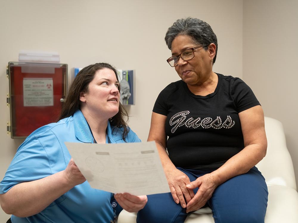 A doctor holds a piece of paper toward a woman patient who is seated on an exam table. The doctor is wearing a blouse with a lanyard around her neck. The woman patient is wearing glasses and a T-shirt with the “Guess” logo on it.