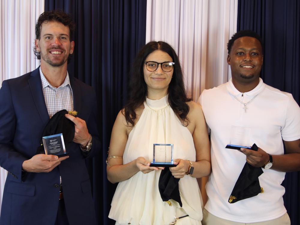 Three students pose for a photo holding clear glass awards in front of a blue and white backdrop.