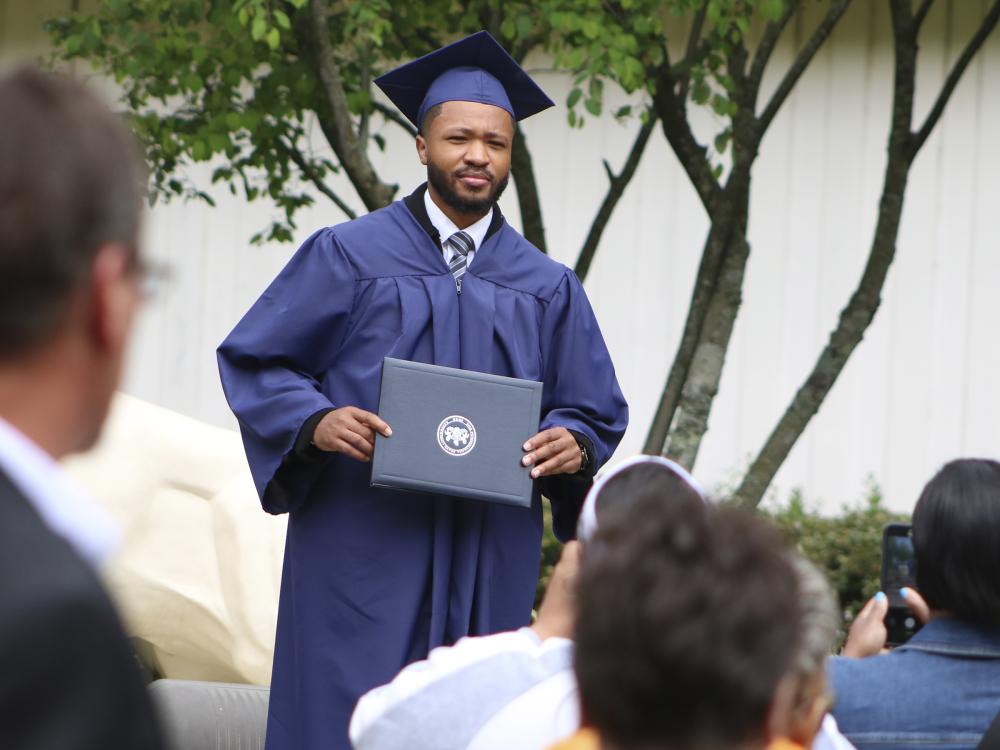 Graduate wearing blue cap and gown poses for a photo holding a diploma with family members blurred in the foreground taking pictures.