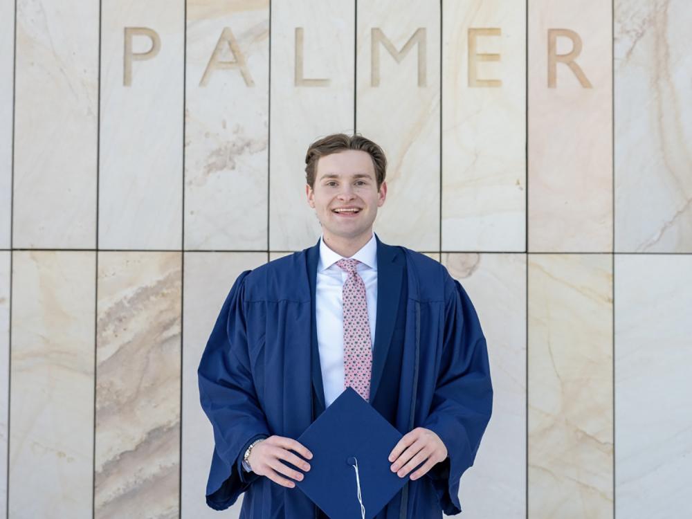 A photo of a smiling Palmer Moore in a commencement gown, holding a graduation cap and standing in front of the words PALMER on a wall behind him.