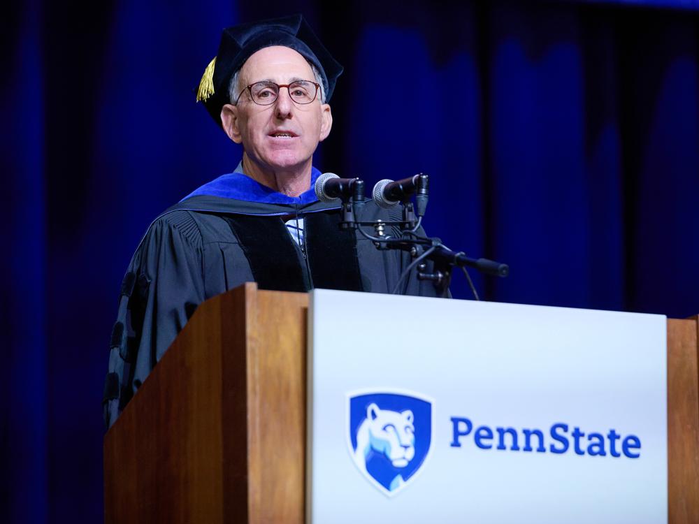 A photo of Stuart Rothstein in a cap and gown, behind a lectern with a Penn State logo speaking at commencement