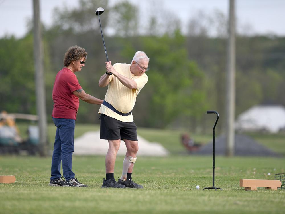 A man swings a golf club while wearing a gait belt and leg brace on his left leg. A man on the left holds his gait belt to give him balance support. A cane is to the right of the golfer.