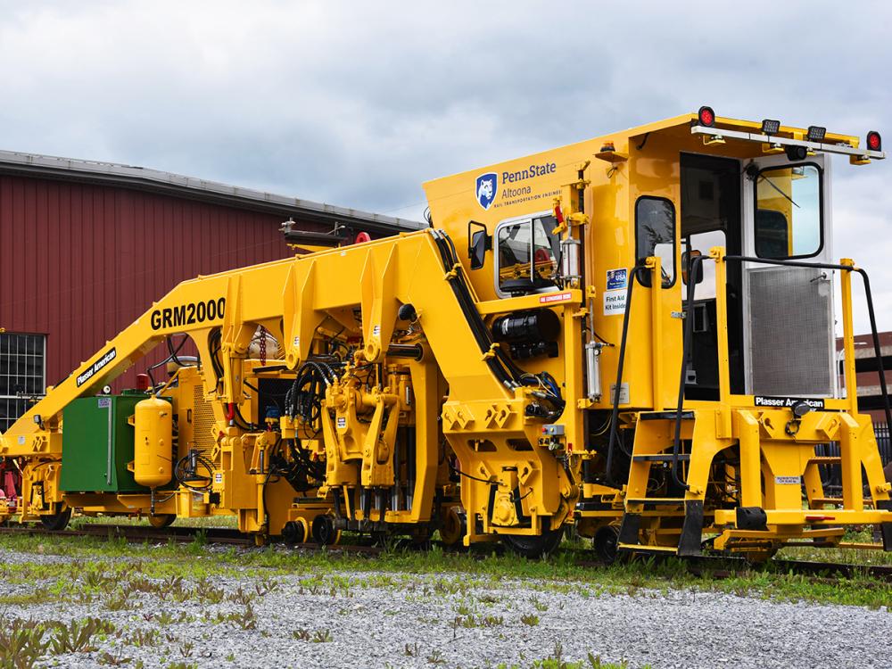 Bright yellow Penn State Altoona GRM2000 rail maintenance machine parked on tracks beside red building.