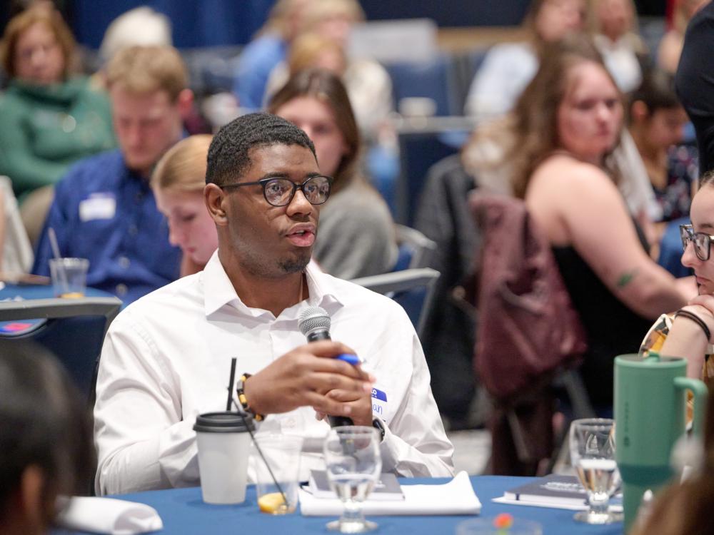 Man in white shirt holding microphone and pen, speaking at a conference with attendees seated around tables.