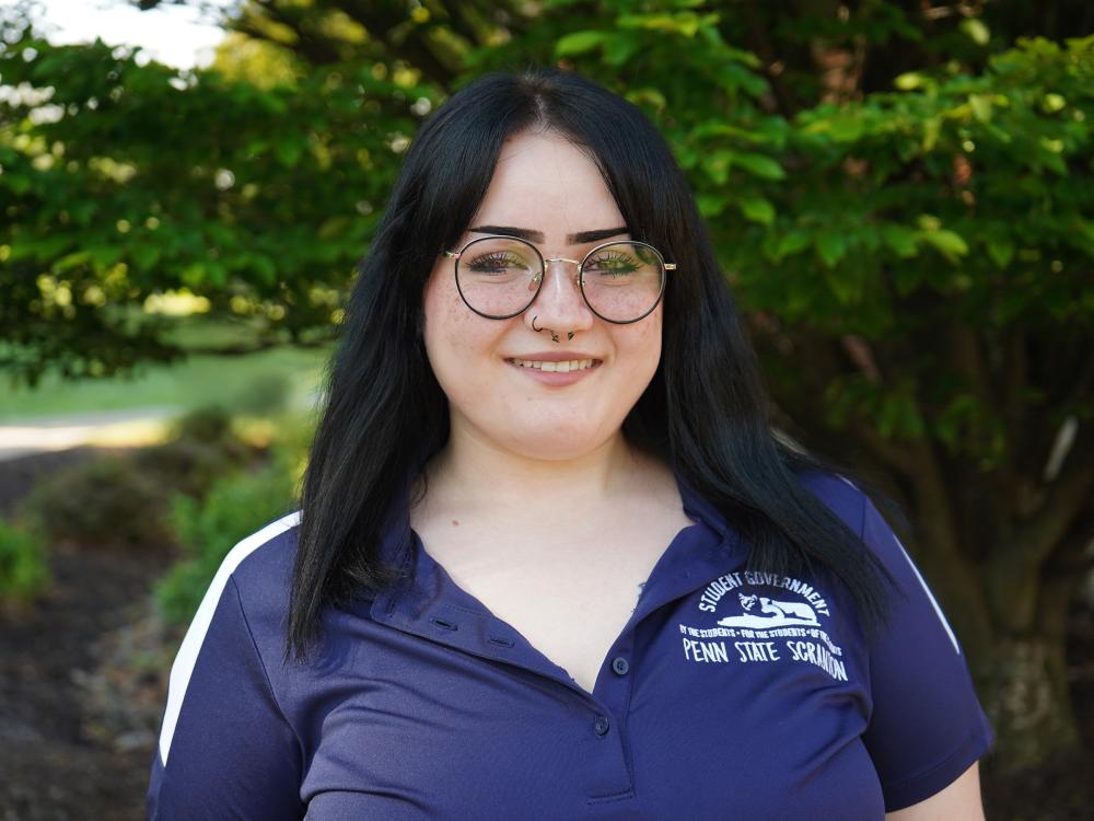 Female student wearing blue and white shirt smiles with trees in background