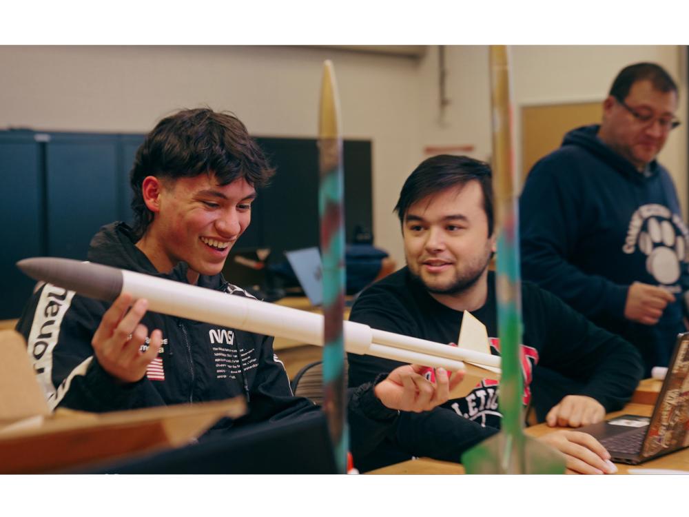 Two students at a table look excited as they work on a model rocket between them