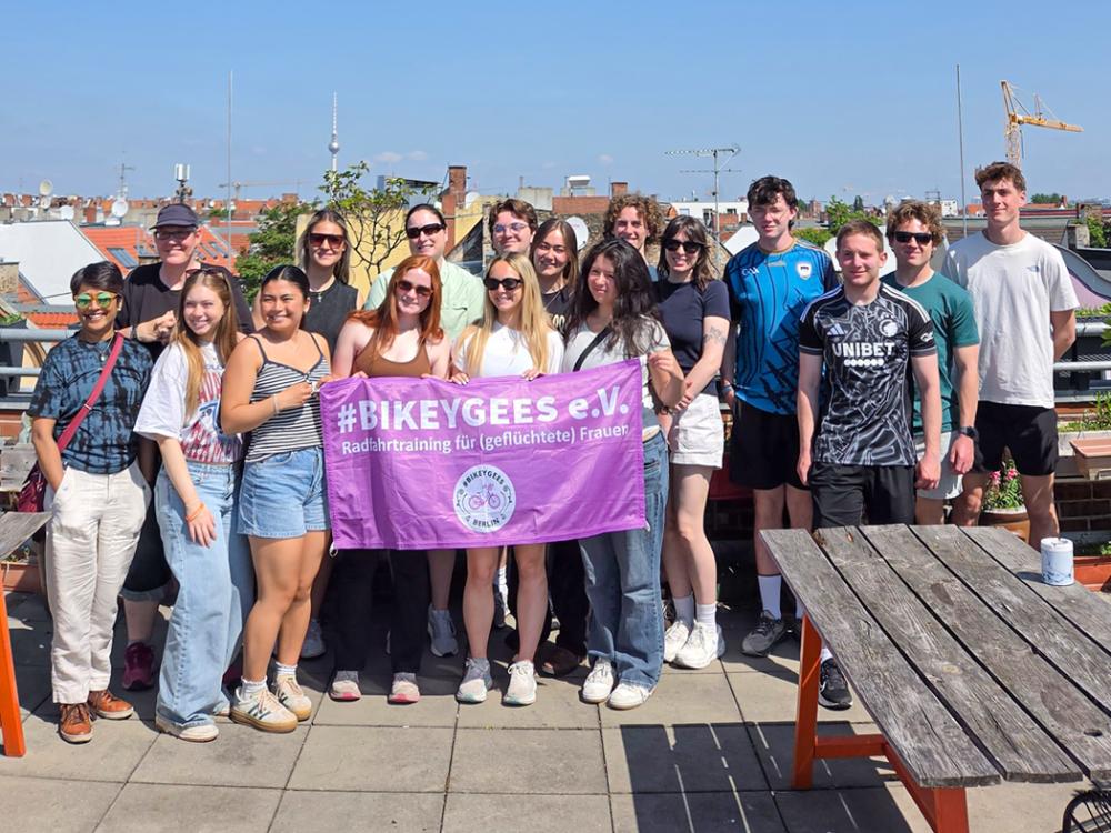 Group of approximately 16 people standing on a rooftop terrace in a city, holding a purple banner that reads “#BIKEYGEES e.V. – Radfahrtraining für (geflüchtete) Frauen.” Buildings and a TV tower are visible in the background under a clear blue sky.