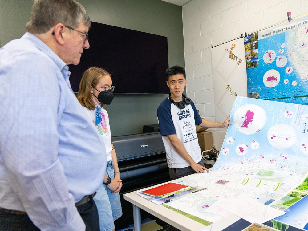 Three people stand around a table looking at large maps and charts. Myers on the left looks at the map while Lily Houtman and Zhaoxu Sui explain the materials.