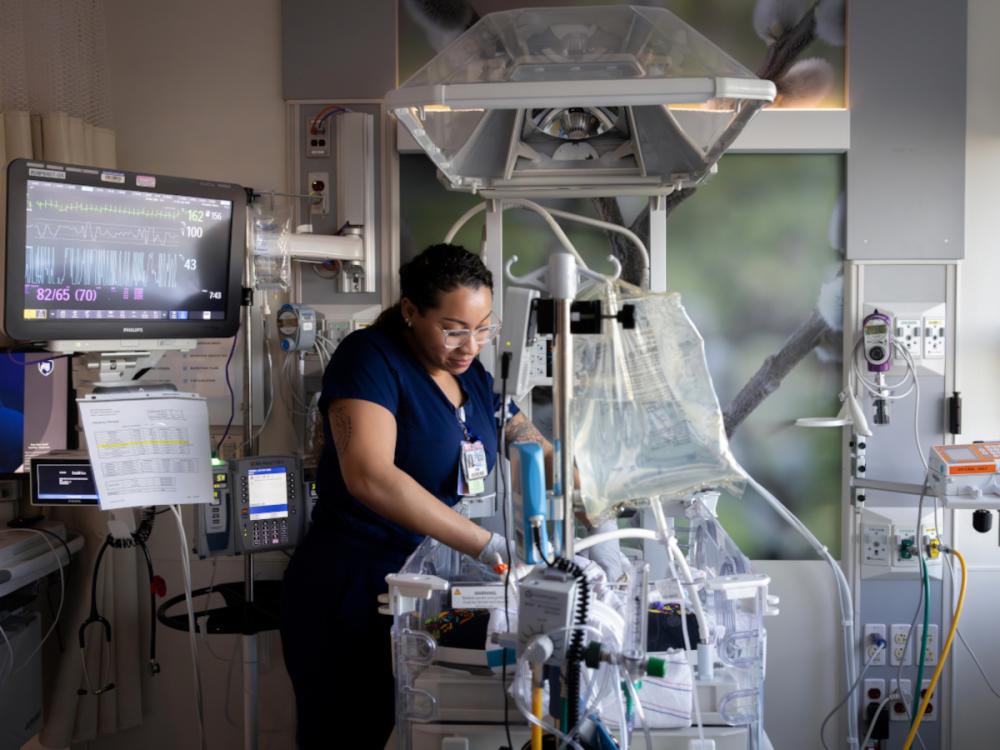 A neonatal intensive care unit (NICU) nurse adjusts equipment and cares for an infant inside an incubator. Medical monitors, IV pumps, and respiratory equipment surround the bedside, while a vital signs monitor displays the baby's heart rate and other readings.