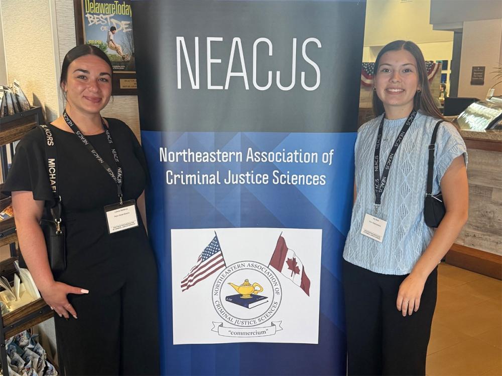 Two women wearing conference badges stand beside a NEACJS banner at a criminal justice sciences event.