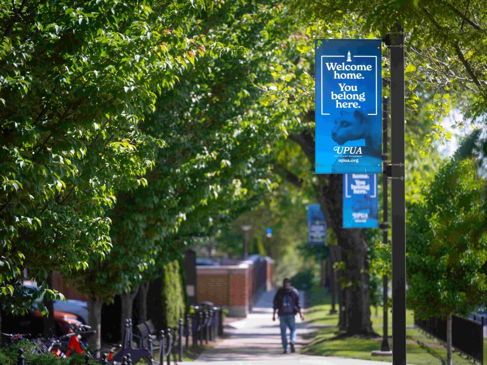A lone student walks away from the camera next to a flag that reads "Welcome home. You belong here."