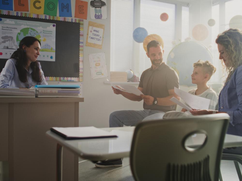 mother, father, approximately 8-year-old boy, and teacher all looking pleased at parent-teacher conference in a classroom