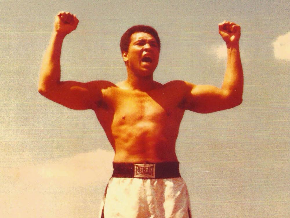 The image shows Muhammad Ali with his arms and fists raised in the air.