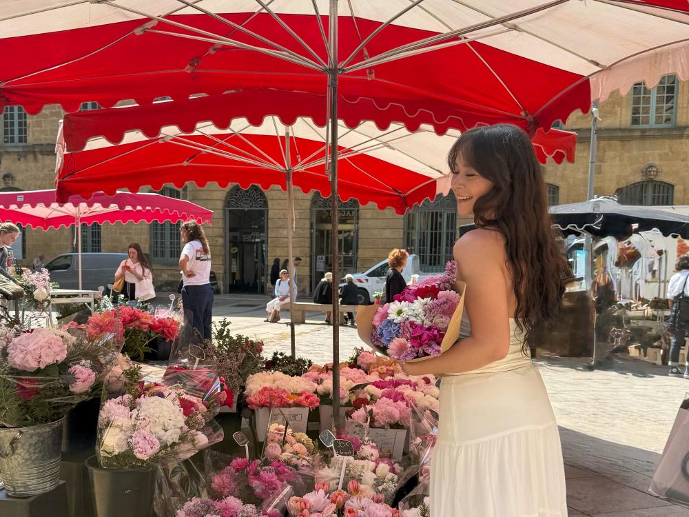 Jenna Guarrera wears a white dress while smiling and holding a bouquet of pink, purple, and white flowers in front of a flower stand on the streets of France.