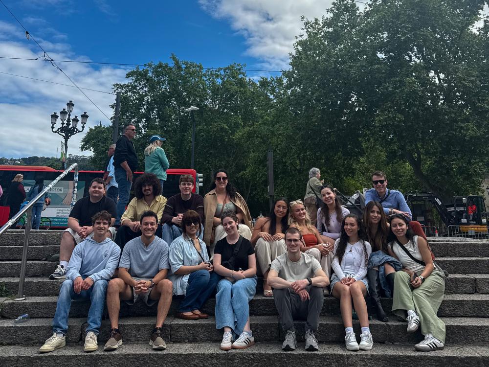 A group of students sit on stairs, smiling at the camera with trees and a red bus in the background.
