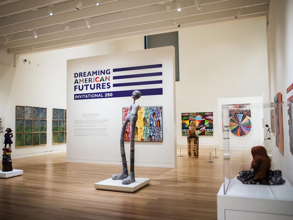 Vibrant and colorful 2D and 3D artworks are on display in a white art museum gallery around a central title wall with a purple flag-motif logo and abstract rainbow artwork.