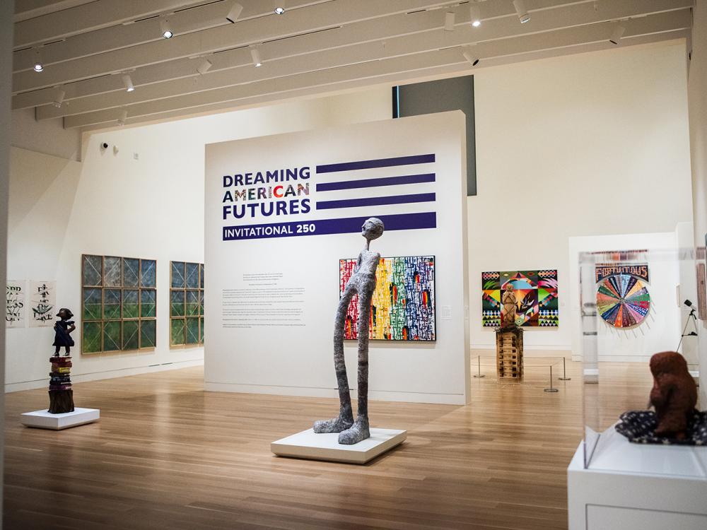 Vibrant and colorful 2D and 3D artworks are on display in a white art museum gallery around a central title wall with a purple flag-motif logo and abstract rainbow artwork.