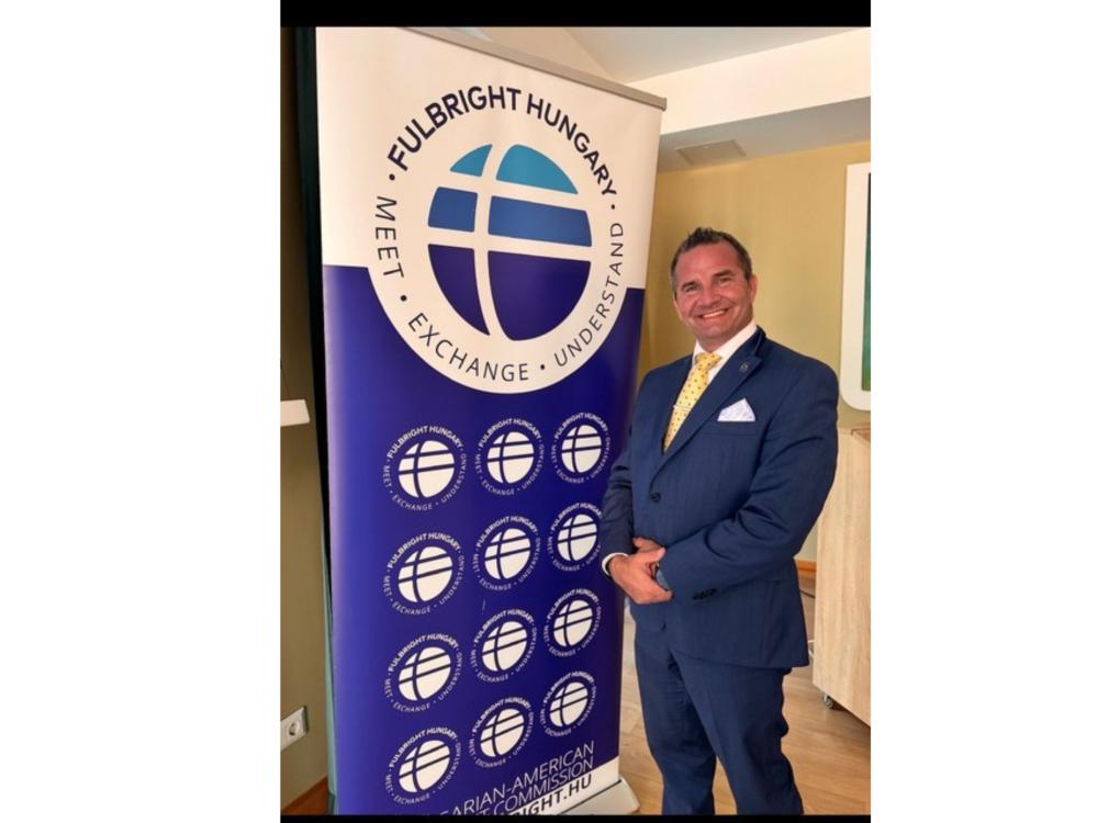 professor in blue suit poses next to Fulbright Hungary sign